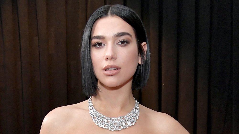 Dua Lipa posing at an event with short hair and a large, sparkly necklace