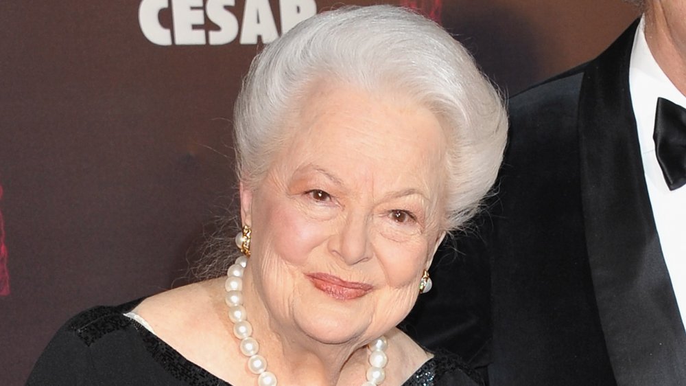 Olivia de Havilland dans une robe noire et des perles, avec un petit sourire lors d'un événement sur le tapis rouge