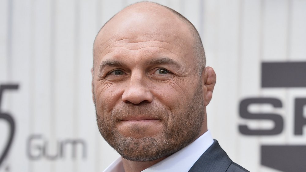 Randy Couture dans un costume gris foncé et chemise blanche, regardant sur le côté avec un petit sourire