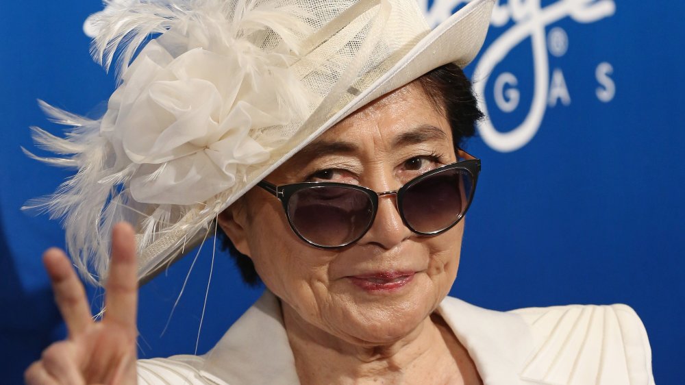 Yoko Ono dans un blazer blanc, chapeau, et lunettes de soleil, sourire et donner le signe de paix