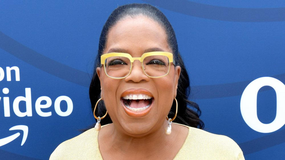 Oprah Winfrey dans une robe jaune et des lunettes, riant tout en regardant directement la caméra