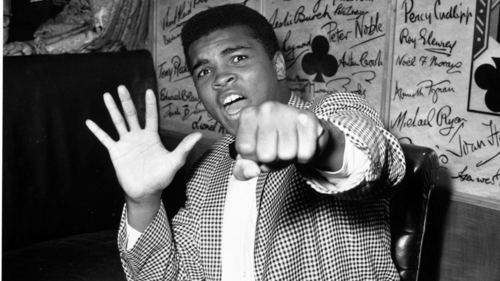 Photo en noir et blanc de Muhammad Ali dans des vêtements de rue, frappant une pose de boxe ludique