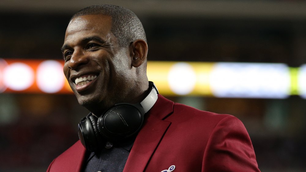 Deion Sanders souriant, tout en regardant sur le côté, dans un blazer marron et un polo noir