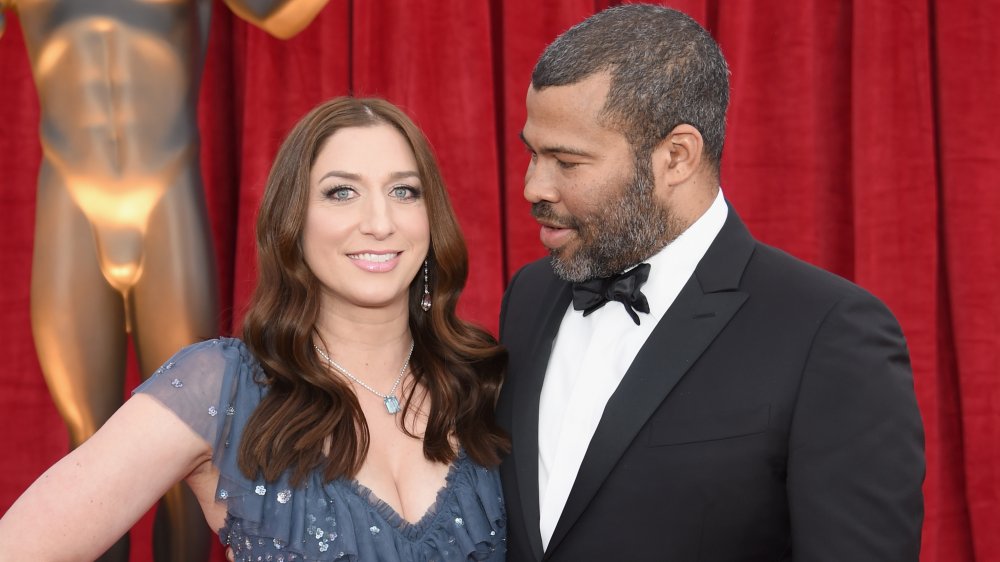 Chelsea Peretti souriant dans une robe bleue, Jordan Peele dans un costume noir regardant son