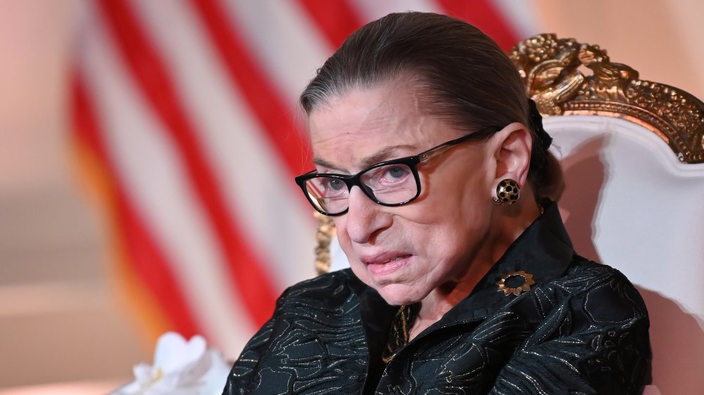 Ruth Bader Ginsburg assise, boucles d’oreilles en or et lunettes noires