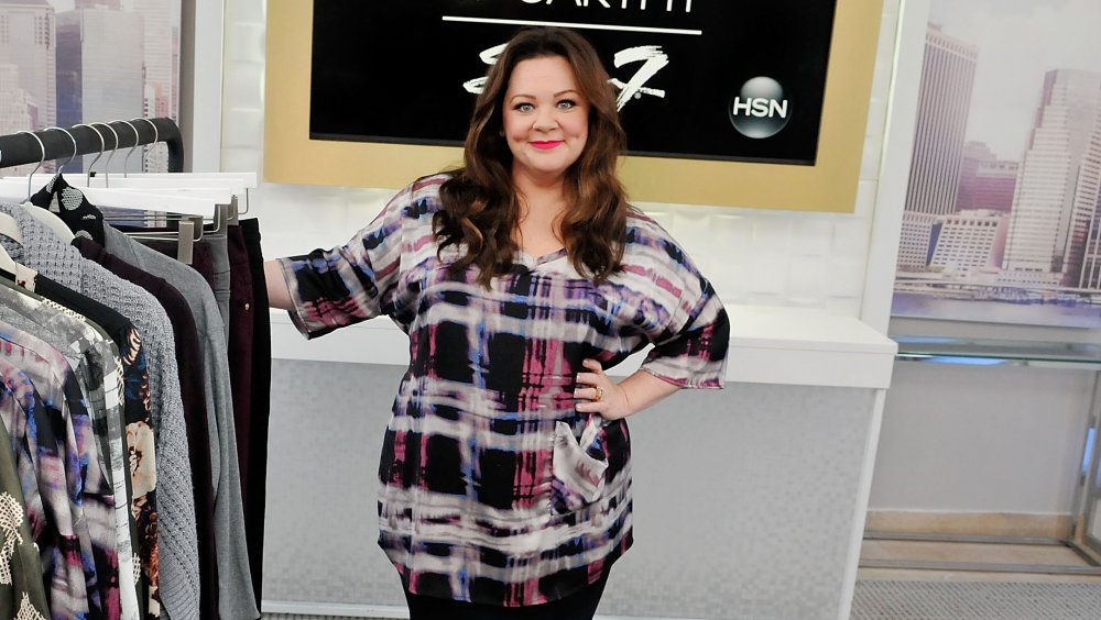 Melissa McCarthy posant à côté d’un porte-vêtements pour sa ligne de vêtements Seven7