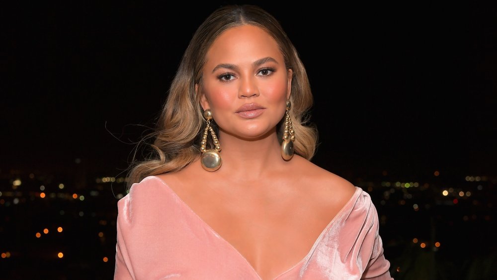 Chrissy Teigen dans une robe rose, posant avec une expression neutre sur un balcon dans un contexte de ville