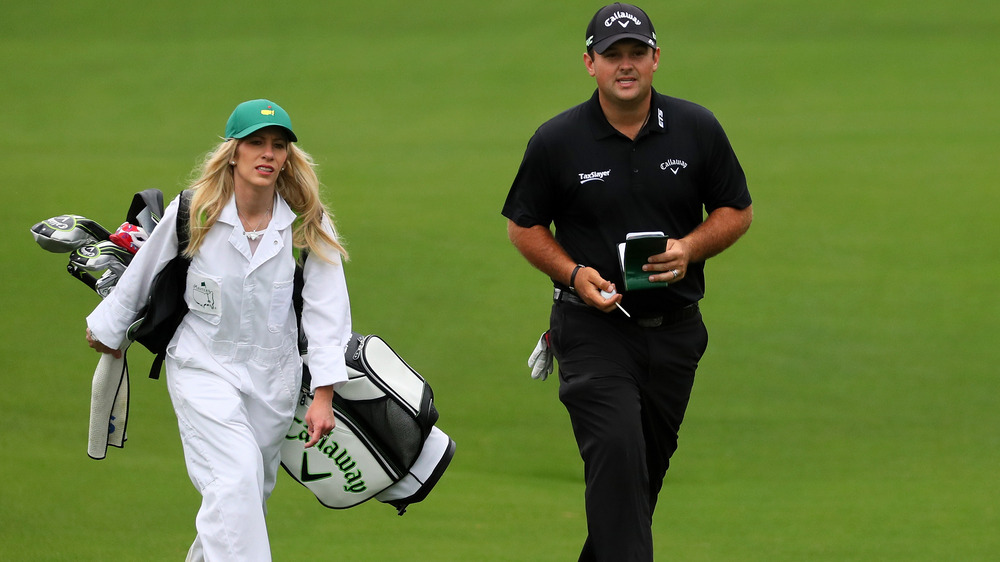 Justine Reed, travaillant comme caddie, vêtue de blanc, portant une haine, tenant des clubs de golf; Patrick Reed, tout noir et un chapeau, sur le terrain de golf