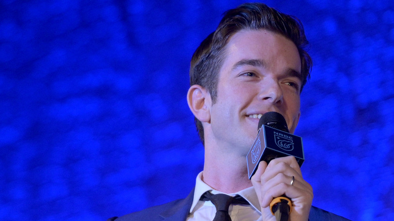 John Mulaney souriant et parlant dans un microphone portant une cravate