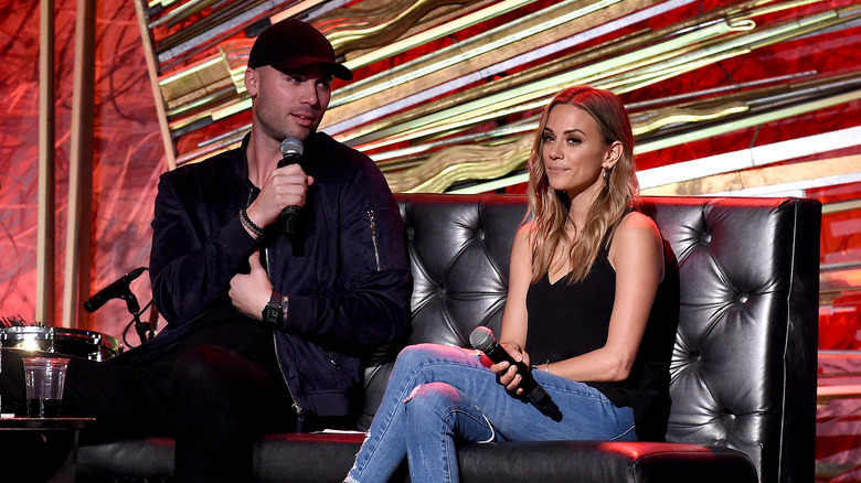 Mike Caussin, vêtu de vêtements sombres et d'un chapeau, parlant dans un micro ; Jana Kramer, assise sur un canapé, l'air agacée, portant un jean, photo de 2019