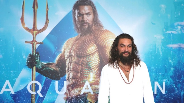 Jason Momoa smiling in front of an Aquaman poster