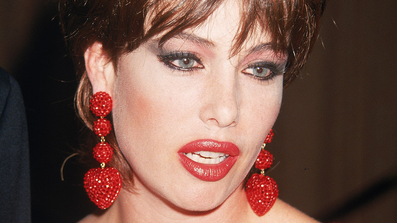 Kelly LeBrock porte du rouge à lèvres rouge et des boucles d'oreilles assorties.