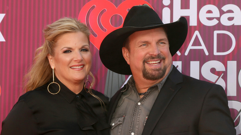 Trisha Yearwood et Garth Brooks sourient sur le tapis rouge tandis que Yearwood porte un chemisier noir et Brooks porte un chapeau de cowboy noir et un blazer assorti