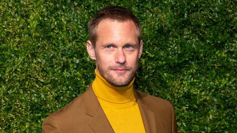 Alexander Skarsgård pose en col roulé jaune et costume marron