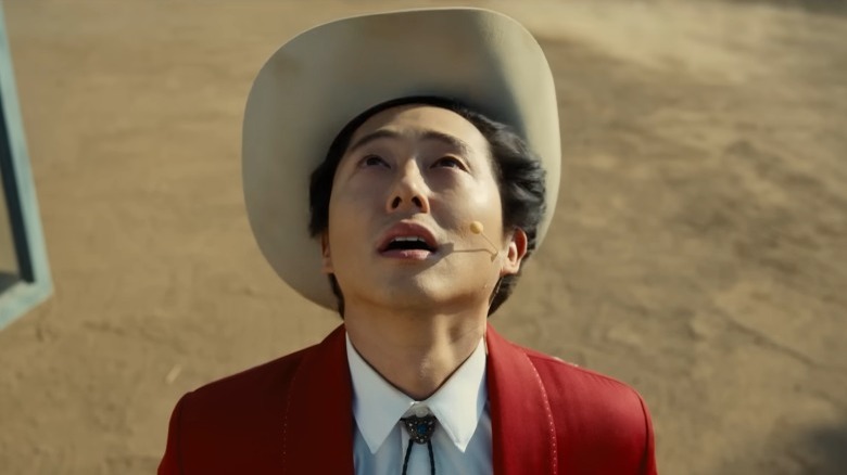 Steven Yeun dans un chapeau de cow-boy, une veste rouge, levant les yeux