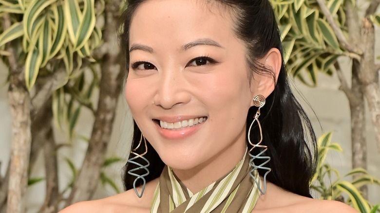 Arden Cho souriant de grosses boucles d'oreilles devant des arbres