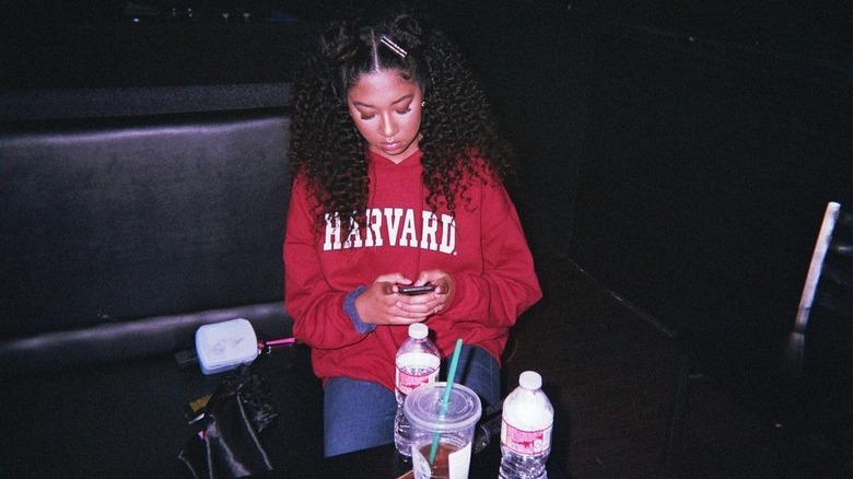 Aoki Lee Simmons sur son téléphone portant un sweat à capuche Harvard