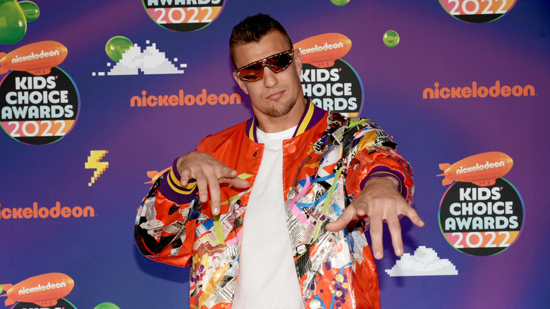 Rob Gronkowski porte une veste orange et à motifs fluo, un t-shirt blanc et des lunettes de soleil foncées tout en pointant vers la caméra