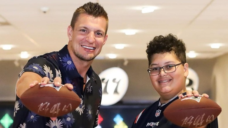 Rob Gronkowski souriant tenant un ballon de football avec un enfant portant des lunettes et un maillot de football des Patriots