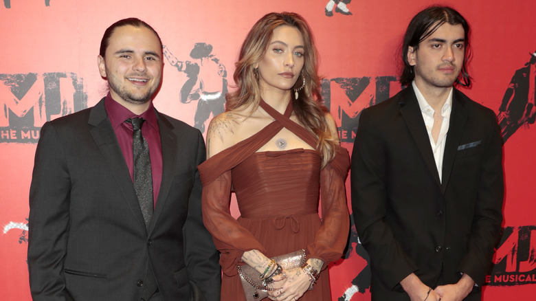 Prince Jackson portant une veste noire, une chemise rouge et une cravate noire ; avec Paris Jackson portant une robe croisée de couleur rouille ; avec Bigi Jackson portant une veste noire et une chemise blanche