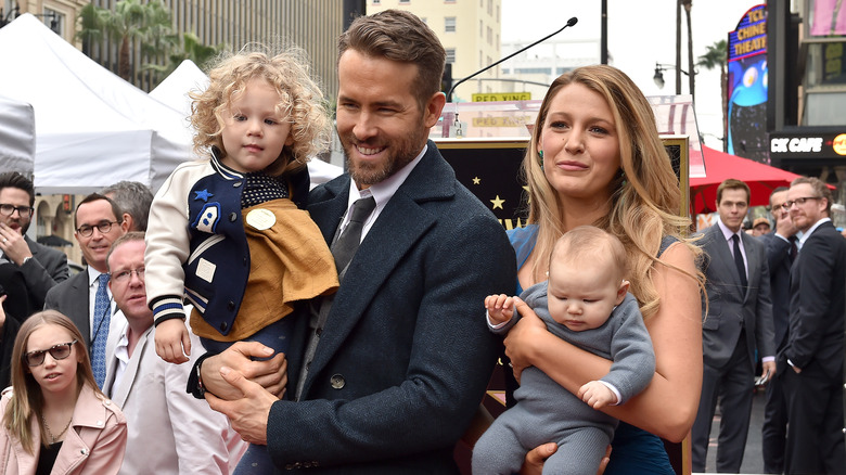 Ryan Reynolds porte un costume noir et une cravate avec Blake Lively dans une robe bleue tandis que tous deux tiennent leurs enfants