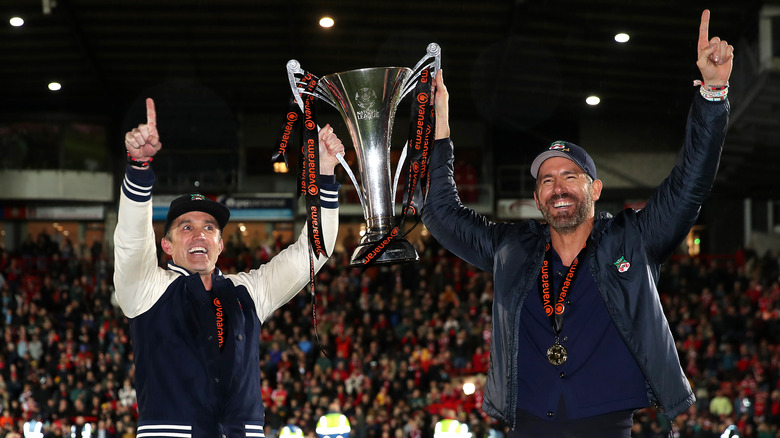 Rob McElhenny et Ryan Reynolds portant l'équipement du Wrexham AFC tout en tenant un trophée