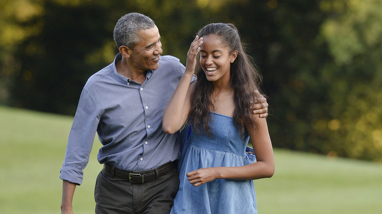 Barack Obama porte une chemise boutonnée grise et un pantalon marron avec Malia Obama portant une robe d'été en jean à bretelles