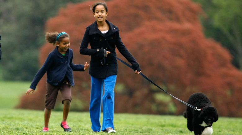 Sasha Obama porte une veste bleue et un short marron avec Malia Obama portant un manteau noir, un jean bleu et tenant un chien en laisse