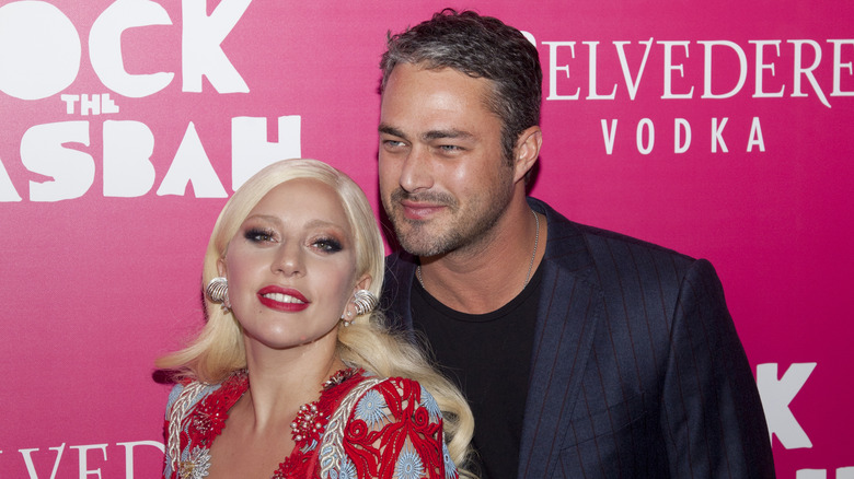 Lady Gaga portant une robe à perles multicolores et Taylor Kinney portant une veste bleue à fines rayures sur le tapis rouge