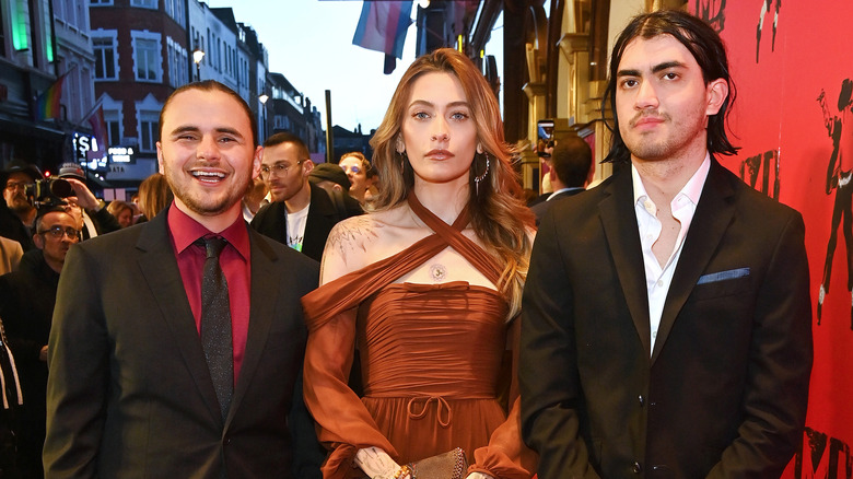 Prince Jackson portant une veste noire, une chemise rouge et une cravate noire ; avec Paris Jackson portant une robe croisée de couleur rouille ; avec Bigi Jackson portant une veste noire et une chemise blanche