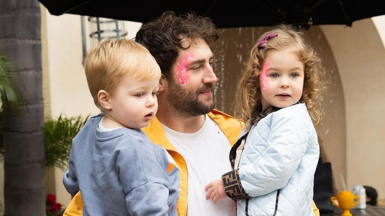 Jordan McGraw tenant ses deux jeunes enfants sur ses hanches tout en portant de la peinture rose pour le visage et une veste jaune