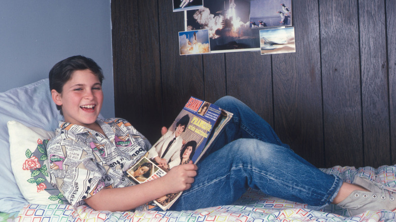 Joaquin Phoenix, enfant, portant une chemise boutonnée à motif journal et un jean, allongé sur un lit