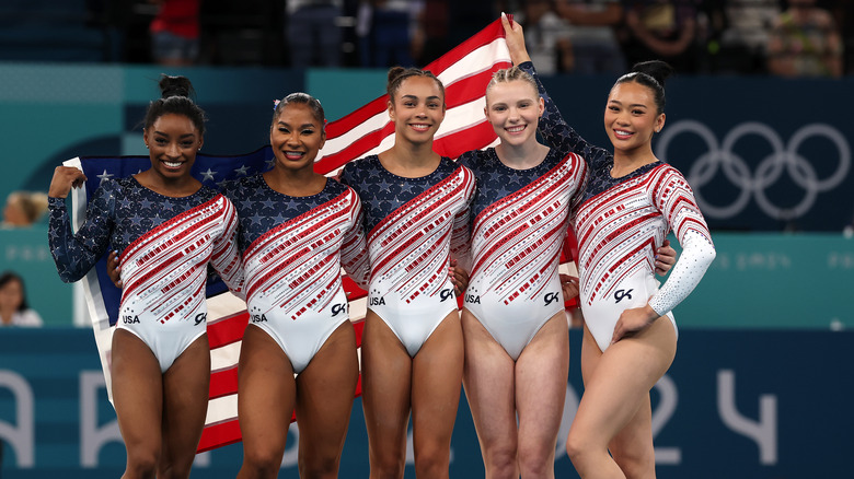 L'équipe olympique féminine américaine de gymnastique de 2024 porte des justaucorps rouges, blancs et bleus étincelants tenant un drapeau américain derrière eux.