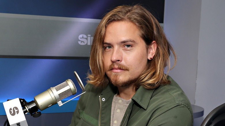 Dylan Sprouse avec un microphone tout en portant un blouson aviateur vert et une chemise boutonnée marron clair