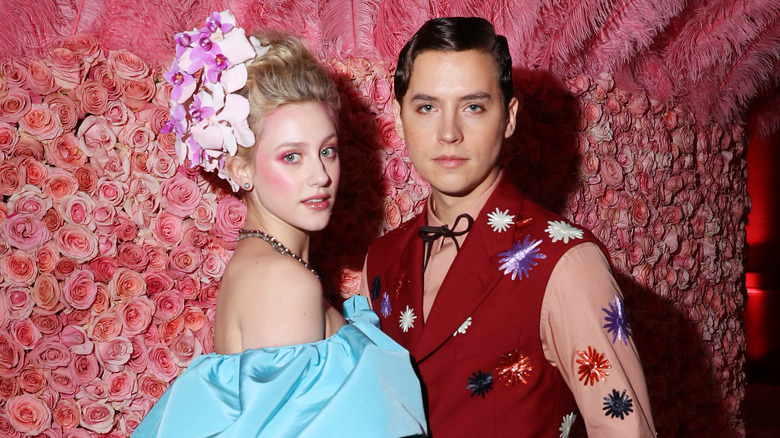 Lili Reinhart dans une perruque fleurie couture et une robe bleue sans manches avec Cole Sprouse portant un gilet fleuri rouge et une chemise à manches longues devant un mur de fleurs