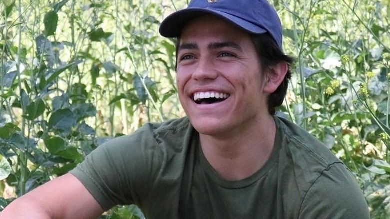 Anthony Barajas souriant assis au milieu de la végétation avec un chapeau bleu et une chemise verte